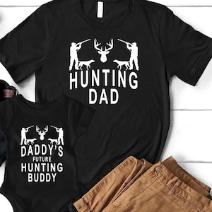 May include: Black t-shirt with the text "Hunting Dad" and a silhouette of a deer and two hunters. A black bodysuit with the text "Daddy's Future Hunting Buddy" and a silhouette of a deer and two hunters.