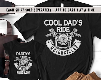May include: Two black t-shirts with a motorcycle graphic. The larger shirt says "Cool Dad's Ride Motorcycles". The smaller shirt says "Daddy's Future Motorcycles Riding Buddy".