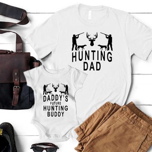 May include: White t-shirt with the text "Hunting Dad" and a silhouette of a deer and two hunters with dogs. A white onesie with the text "Daddy's Future Hunting Buddy" and a similar silhouette of a deer and two hunters with dogs.