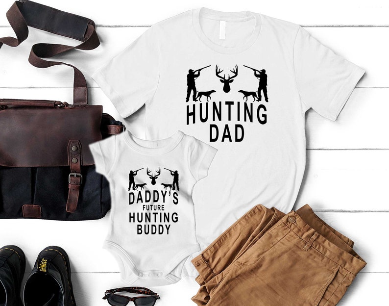 May include: White t-shirt with the text "Hunting Dad" and a silhouette of a deer and two hunters with dogs. A white onesie with the text "Daddy's Future Hunting Buddy" and a similar silhouette of a deer and two hunters with dogs.
