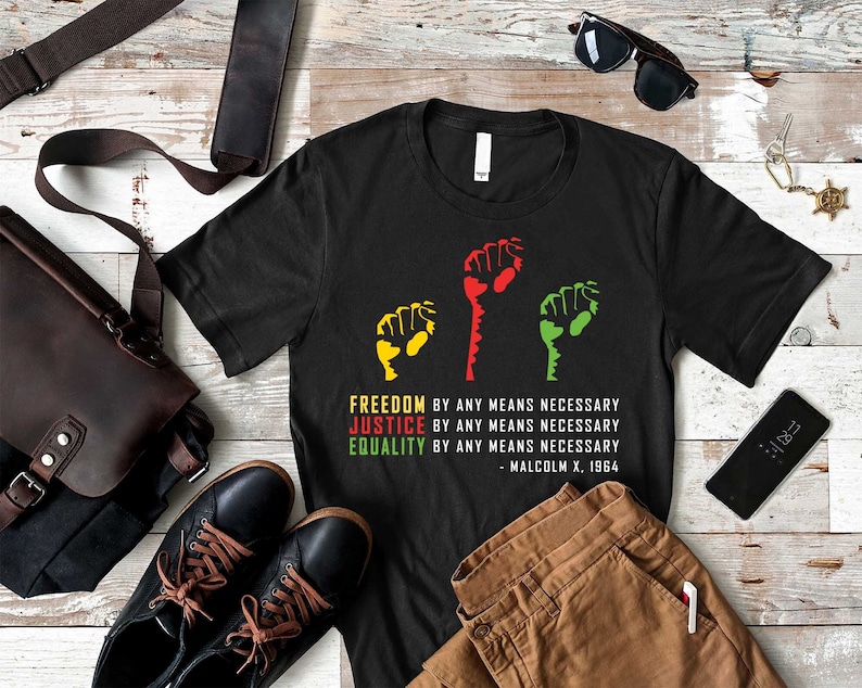 May include: Black t-shirt with a graphic of three raised fists in red, yellow, and green. The text "FREEDOM BY ANY MEANS NECESSARY, JUSTICE BY ANY MEANS NECESSARY, EQUALITY BY ANY MEANS NECESSARY - MALCOLM X. 1964" is printed below the fists.