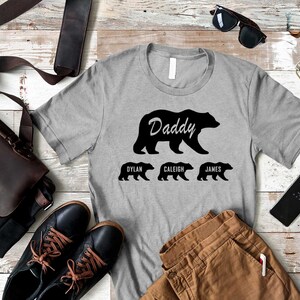 K&ouml;nnte beinhalten: Ein graues T-Shirt mit einer schwarzen B&auml;rensilhouette und dem Wort "Daddy" dar&uuml;ber. Drei kleinere B&auml;rensilhouetten befinden sich unter dem gro&szlig;en B&auml;ren, mit den Namen "Dylan", "Caleigh" und "James" darunter.