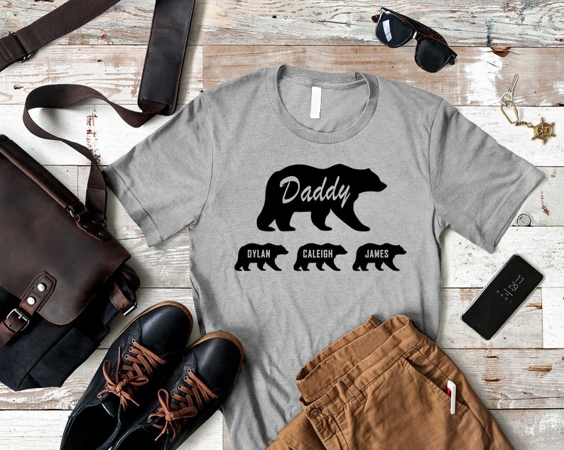 K&ouml;nnte beinhalten: Ein graues T-Shirt mit einer schwarzen B&auml;rensilhouette und dem Wort "Daddy" dar&uuml;ber. Drei kleinere B&auml;rensilhouetten befinden sich unter dem gro&szlig;en B&auml;ren, mit den Namen "Dylan", "Caleigh" und "James" darunter.