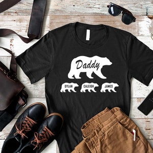 K&ouml;nnte beinhalten: Schwarzes T-Shirt mit einer wei&szlig;en B&auml;rensilhouette und dem Wort "Daddy" im B&auml;ren. Drei kleinere B&auml;rensilhouetten befinden sich unter dem gro&szlig;en B&auml;ren mit den Namen "Dylan", "Caleig" und "James" darunter.