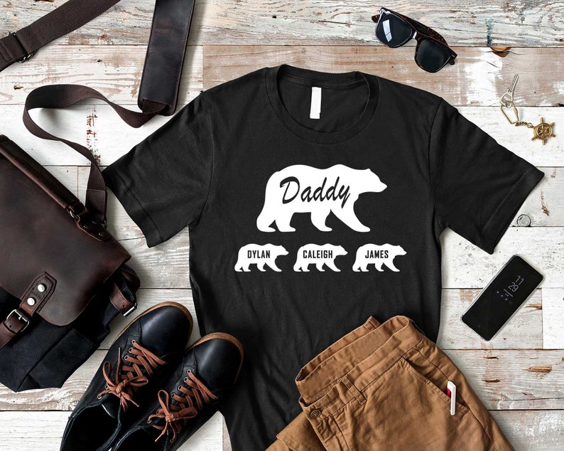K&ouml;nnte beinhalten: Schwarzes T-Shirt mit einer wei&szlig;en B&auml;rensilhouette und dem Wort "Daddy" im B&auml;ren. Drei kleinere B&auml;rensilhouetten befinden sich unter dem gro&szlig;en B&auml;ren mit den Namen "Dylan", "Caleig" und "James" darunter.