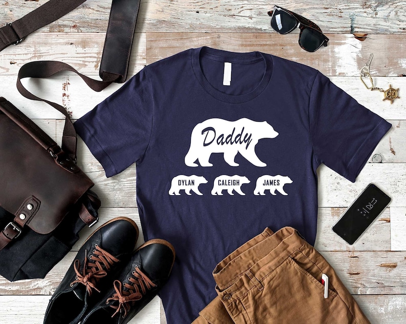 K&ouml;nnte beinhalten: Ein marineblaues T-Shirt mit einer wei&szlig;en B&auml;rensilhouette und dem Text "Daddy" dar&uuml;ber. Drei kleinere B&auml;rensilhouetten befinden sich unter dem gro&szlig;en B&auml;ren mit den Namen "Dylan", "Caleigh" und "James" darunter.
