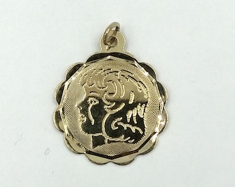 May include: Gold pendant charm featuring a detailed profile of a person's face with a scalloped edge.
