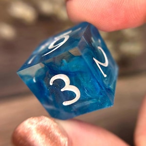 May include: A blue and white D20 dice with white numbers. The dice is held between two fingers. The dice has a translucent blue base with white swirls. The numbers 2, 3, and 9 are visible. The dice is for tabletop games.