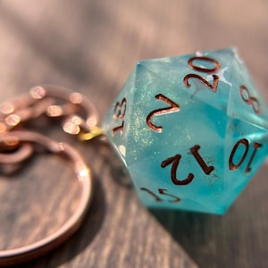 May include: A keychain featuring a translucent light blue twenty-sided die with gold glitter flecks embedded inside. The die has copper-colored numbers engraved on its faces and is attached to a circular copper-toned metal split ring.