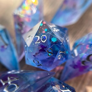 May include: A set of polyhedral dice made of translucent blue and purple resin with swirling patterns, embedded with small butterfly shapes and glitter, featuring silver-colored numbers and a prominent 20 on the central twenty-sided die.