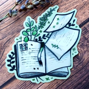 May include: A sticker featuring an open book with pages and botanical elements. The book has a green ribbon bookmark and is surrounded by green leaves and sprigs. The sticker has a light green border and is set against a wooden background.