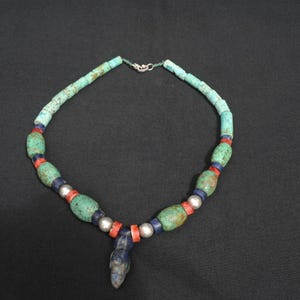 May include: A turquoise beaded necklace with a silver clasp. The necklace features a variety of beads, including turquoise, red, blue, and silver. A blue pendant hangs from the center.