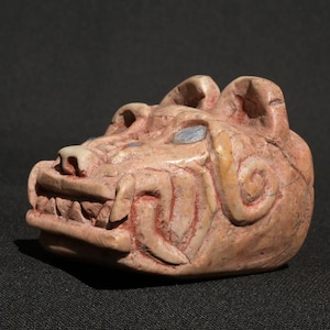 May include: A carved stone sculpture of a snarling animal head, possibly a jaguar or similar feline. The sculpture is a light tan color with red accents in the mouth and around the eyes. The details include sharp teeth and curled ear shapes.