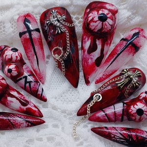 May include: A collection of long, pointed false nails with a dark red base and various designs. Some nails feature a cross, others have an eyeball design, and some have a chain and cross charm. The nails are decorated with black and white accents.
