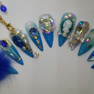 May include: A set of ten blue and white ombre stiletto nails with various gold and crystal embellishments. The nails feature a variety of designs, including a cameo, a key, a crown, and a pearl.