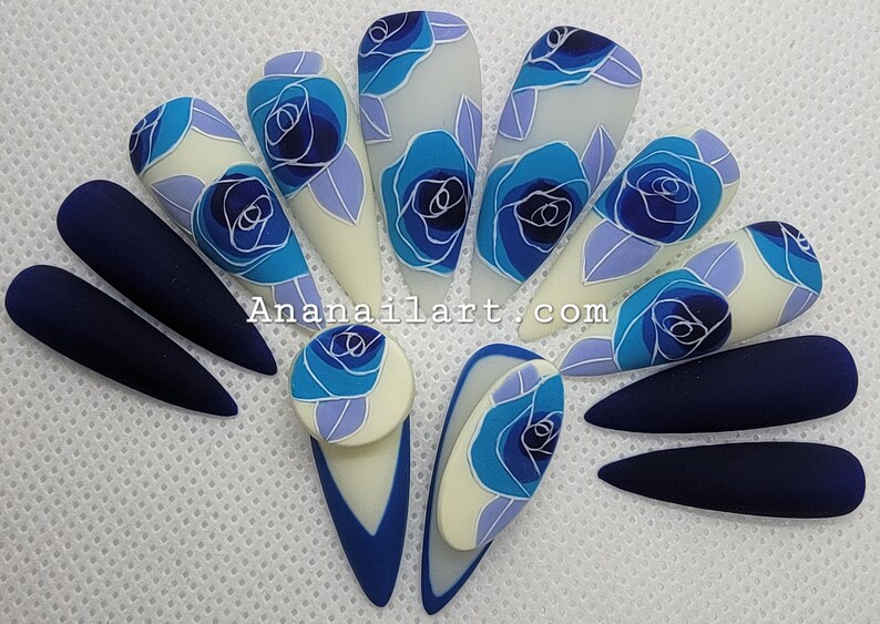 Pu&ograve; includere: Un set di dieci unghie finte lunghe a forma di mandorla con un design floreale blu e bianco. Alcune unghie sono di un blu navy solido, mentre altre presentano una base bianca con rose blu e foglie.