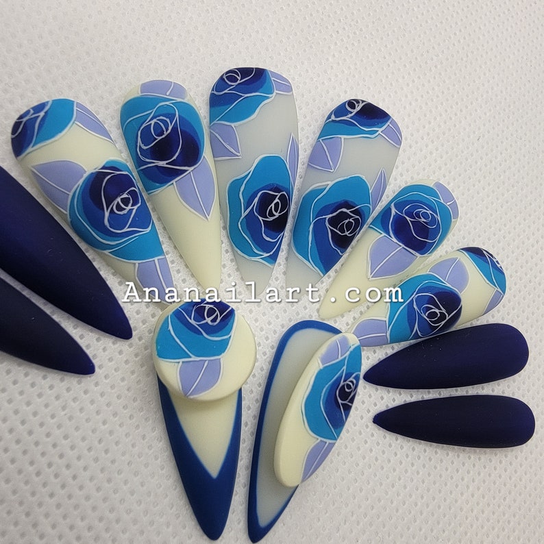 Pu&ograve; includere: Nail art blu e bianca con un design floreale. Le unghie sono lunghe e a forma di mandorla. I fiori sono dipinti in uno stile realistico con petali blu e centri bianchi. Le unghie sono anche decorate con linee e punti bianchi.