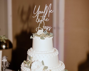 May include: A three-tiered white wedding cake decorated with pearl-like accents and greenery. The top tier features a white flower and a white sign that reads "You Me & the Dogs".