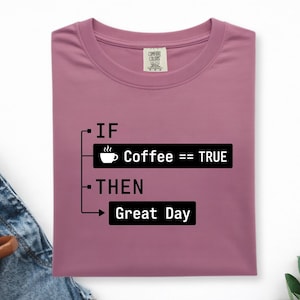 May include: A mauve t-shirt featuring black text and graphics that read: IF Coffee == TRUE THEN Great Day. The design includes a small coffee cup icon and a programming-style logic flow structure printed on the centre of the chest area.
