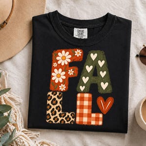 May include: A black t-shirt with the word FALL printed in large block letters. The F is orange with white daisies, the A is olive green with white hearts, the L is leopard print, and the second L is orange plaid with a red heart.