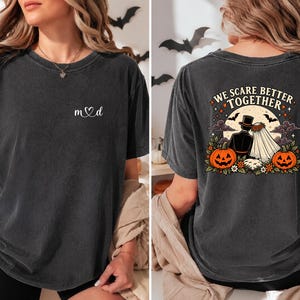 Puede incluir: Una camiseta gris carbón con un pequeño logotipo de corazón blanco en el pecho. La parte posterior muestra un gráfico de una novia y un novio con ropa de boda, dos calabazas naranjas y el texto "WE SCARE BETTER TOGETHER" bajo una luna llena.