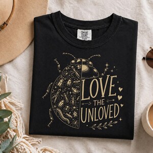 Peut inclure: Un t-shirt noir avec un motif doré représentant un scarabée divisé en deux, une moitié montrant un motif détaillé de divers insectes et l'autre une silhouette unie. Le texte indique LOVE THE UNLOVED, avec de petits cœurs et des étoiles.