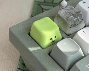 May include: Close-up of a computer keyboard with custom keycaps. One keycap is a light green cube with a simple face, and another is a gray cat. The other keys are white and gray.