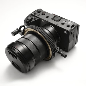 May include: A black digital camera with a lens attached, featuring a lens cap. The camera body has several buttons and dials. The lens has a ribbed design with a gold-colored ring. The camera is labeled "Cinema Line FX30".
