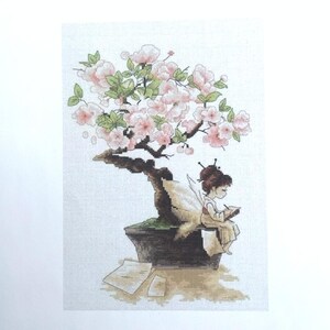 May include: A cross-stitch pattern depicting a fairy seated on a bonsai tree with pink blossoms. The fairy is holding a book and has a pair of wings.