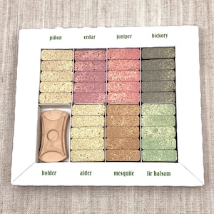 Puede incluir: Una caja blanca con un soporte de madera y 10 filas de varillas de incienso. Las varillas de incienso están dispuestas en una cuadrícula y están etiquetadas con los siguientes aromas: piñon, cedro, enebro, nogal, aliso, mezquite, bálsamo de abeto y soporte.