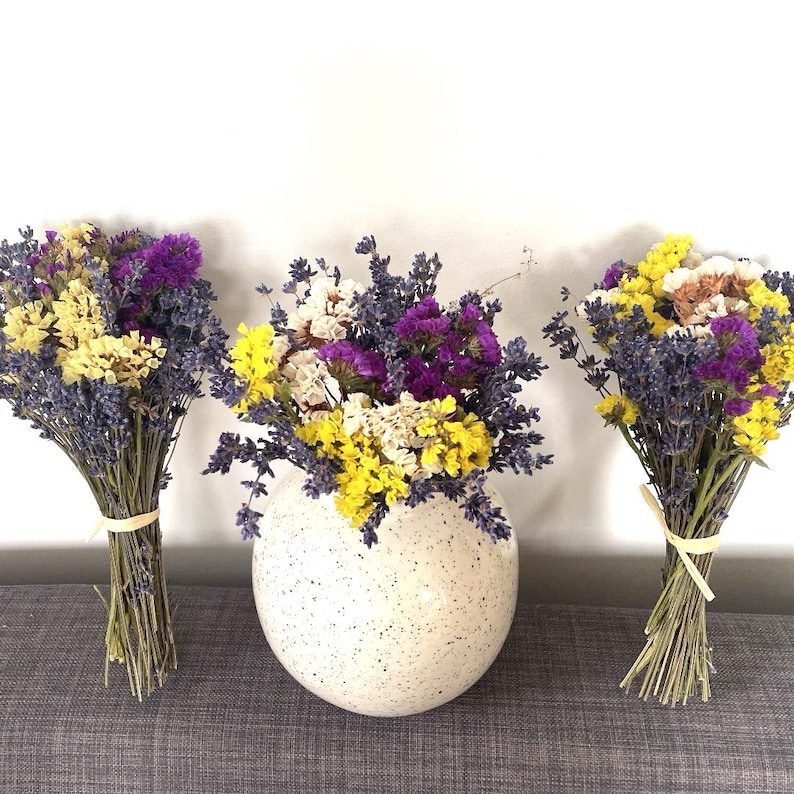 May include: Three bouquets of dried flowers in shades of purple, yellow, and white. The bouquets are tied with twine and arranged in a row on a gray surface. The center bouquet is in a white speckled vase.