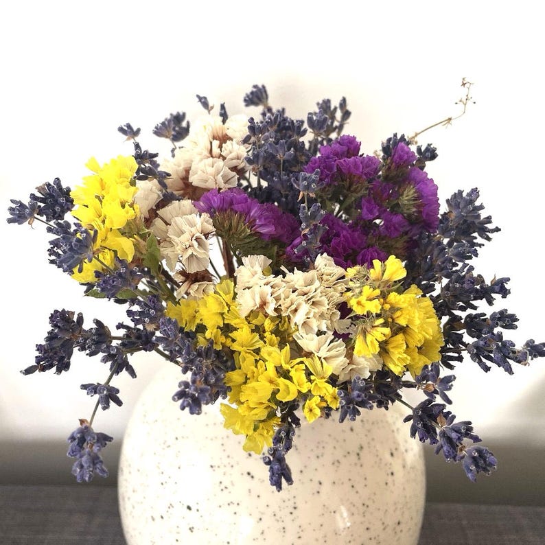 May include: A bouquet of dried flowers in a white speckled vase. The bouquet includes purple lavender, yellow statice, and white baby's breath.