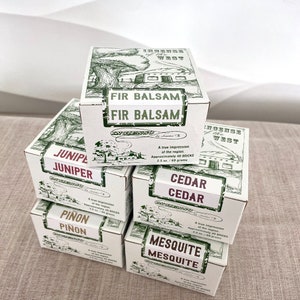 May include: A stack of six white boxes with green illustrations of trees and text. The boxes are labelled with different scents: Fir Balsam, Juniper, Cedar, Piñon, and Mesquite. Each box has the text "Incense of the West" and "Angleinso de Santa Fe".