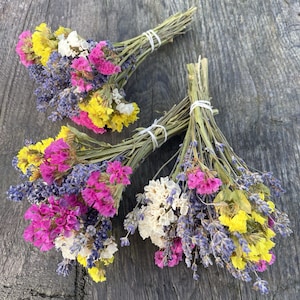 Peut inclure: Trois bouquets de fleurs séchées, chacun lié avec un ruban blanc. Les bouquets présentent un mélange de fleurs de lavande, jaunes, roses et blanches.