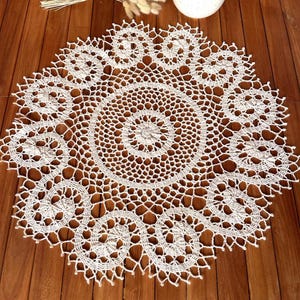 Peut inclure: Une nappe en crochet blanche avec un motif floral délicat. La nappe est circulaire et a un centre avec un cercle plus petit.