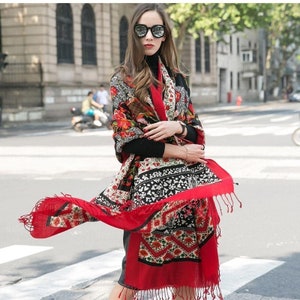 Puede incluir: Una bufanda de patrones rojos, negros y blancos con diseños florales y geométricos. La bufanda tiene flecos en los bordes y está drapeada sobre los hombros de una persona.