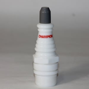 May include: White ceramic spark plug shaped bottle with a gray cap. The bottle has the word "CHAMPION" printed in red on the side.