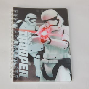 May include: A spiral bound notebook with a black background and a white Stormtrooper from Star Wars. The Stormtrooper is holding a blaster and has a red laser beam coming out of it. The text "STORM TROOPER" is printed on the cover.
