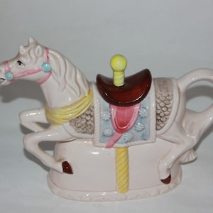 May include: A white ceramic teapot shaped like a carousel horse. The horse has a yellow saddle, pink and blue trim, and a brown saddle. The teapot has a brown handle and a yellow and brown pole.