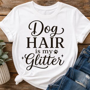 Puede incluir: Una camiseta blanca de manga corta con el texto en negro "Dog HAIR is my Glitter", junto a una peque&ntilde;a huella de pata y estrellas. La prenda cuenta con un cuello redondo y un nudo en el dobladillo inferior, mostrando un dise&ntilde;o sencillo.