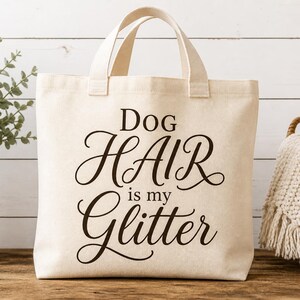 Puede incluir: Un bolso de mano de lona color crema con dos asas superiores. La parte frontal del bolso presenta un texto de color marr&oacute;n oscuro que dice Dog HAIR is my Glitter en una combinaci&oacute;n de fuentes de bloque y cursiva de tela resistente.
