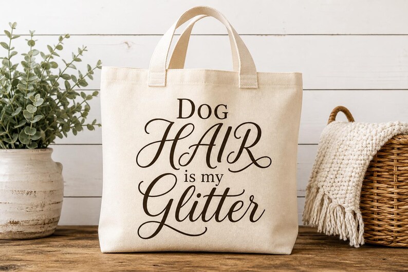 Puede incluir: Un bolso de mano de lona color crema con dos asas superiores. La parte frontal del bolso presenta un texto de color marr&oacute;n oscuro que dice Dog HAIR is my Glitter en una combinaci&oacute;n de fuentes de bloque y cursiva de tela resistente.