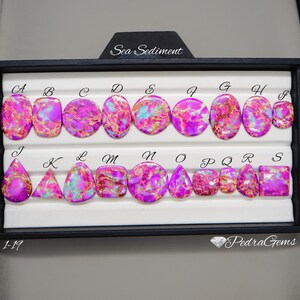 May include: A collection of polished sea sediment gemstones in various shapes, including ovals, circles, and squares. The stones feature a vibrant mix of pink, purple, and turquoise hues, with unique patterns. The gemstones are displayed in a black tray.