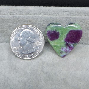 May include: A heart-shaped gemstone with a green and purple pattern. A U.S. quarter is next to the gemstone for size comparison. The gemstone has a polished surface and is set against a gray background.