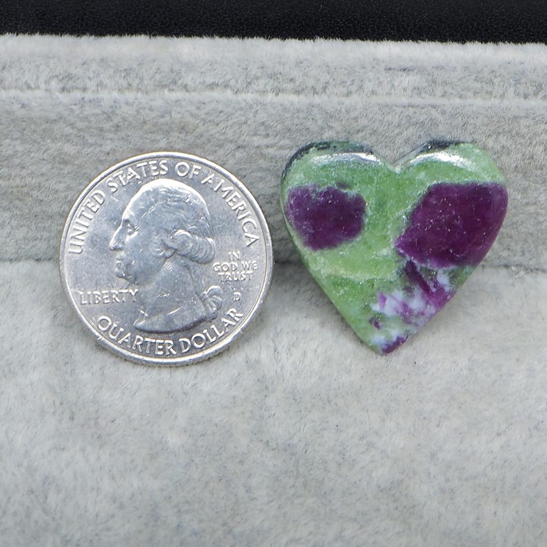 May include: A heart-shaped gemstone with a green and purple pattern. A U.S. quarter is next to the gemstone for size comparison. The gemstone has a polished surface and is set against a gray background.