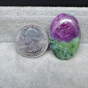 May include: A polished, oval-shaped gemstone with a vibrant color gradient. The top half is a deep magenta, transitioning to a green base. A US quarter coin is next to the gemstone for size comparison.