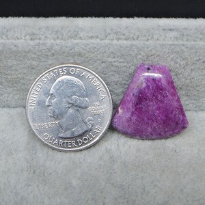 May include: A vibrant, triangular-shaped, polished purple gemstone pendant with a small hole at the top. A US quarter coin is next to the pendant for size comparison. The gemstone has a textured surface and varying shades of purple.