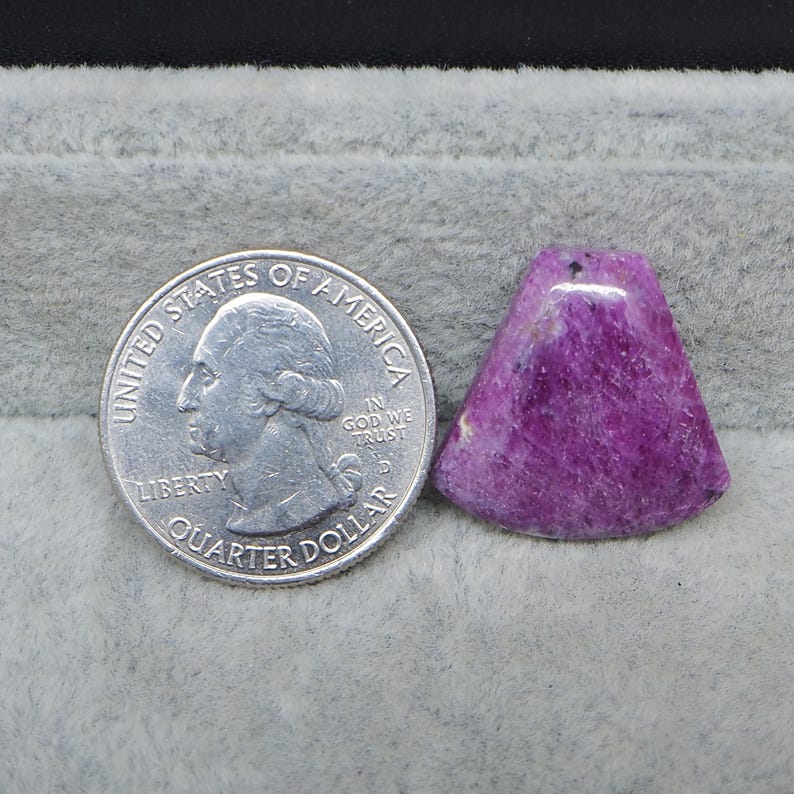 May include: A vibrant, triangular-shaped, polished purple gemstone pendant with a small hole at the top. A US quarter coin is next to the pendant for size comparison. The gemstone has a textured surface and varying shades of purple.