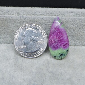 May include: A teardrop-shaped gemstone with a vibrant magenta hue, accented by bands of green and black. A U.S. quarter coin is placed next to the gemstone for size comparison. The stone is set against a gray background.