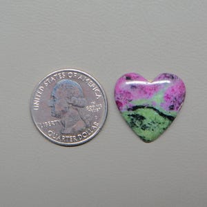 May include: A heart-shaped gemstone with a quarter coin next to it for size comparison. The gemstone features a mix of pink, green, and black colors. The coin is a United States quarter dollar.